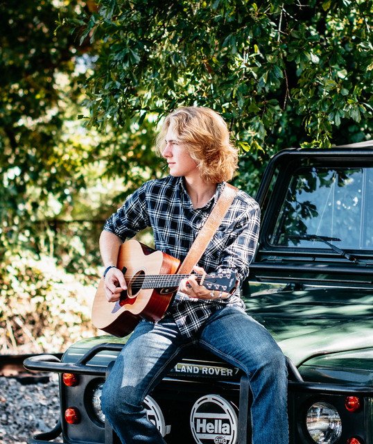Jake Evans seated on the hood of a truck with an acoustic guitar, looking off to one side in dappled afternoon light.