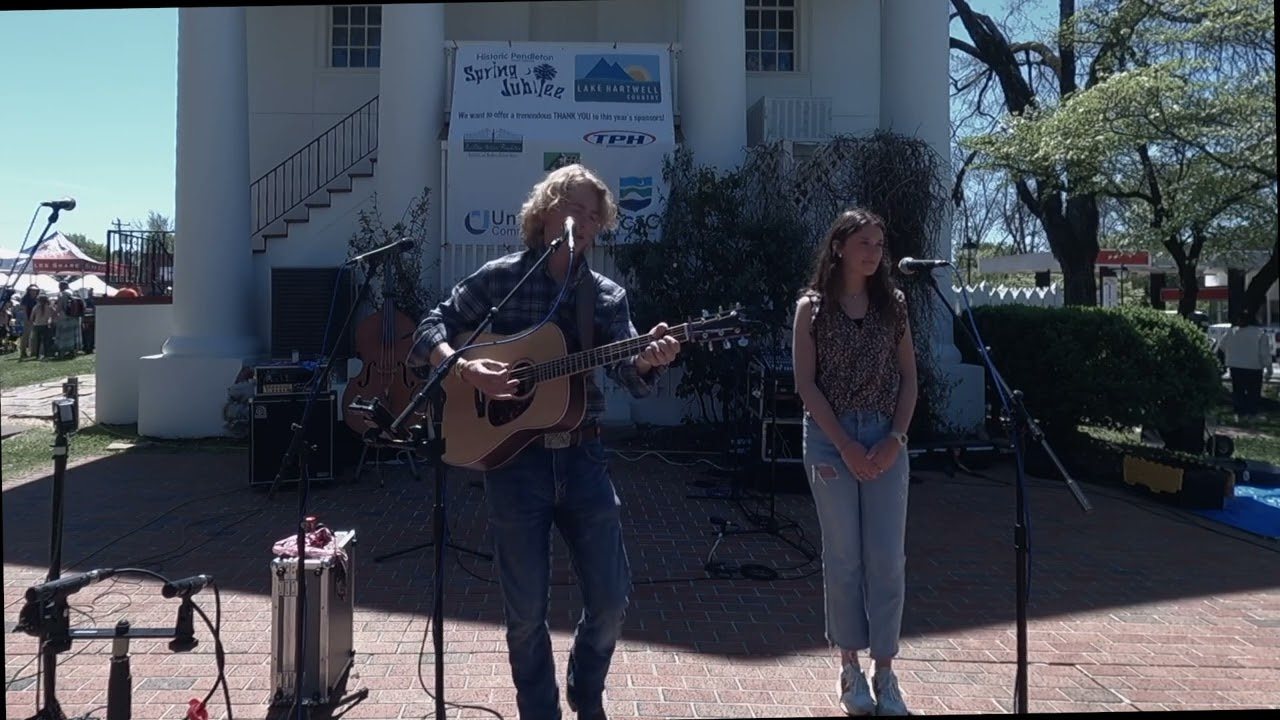Jake Evans playing acoustic guitar and singing into a microphone on a brick plaza outside a white-columned building, with a second performer standing at a microphone beside him.